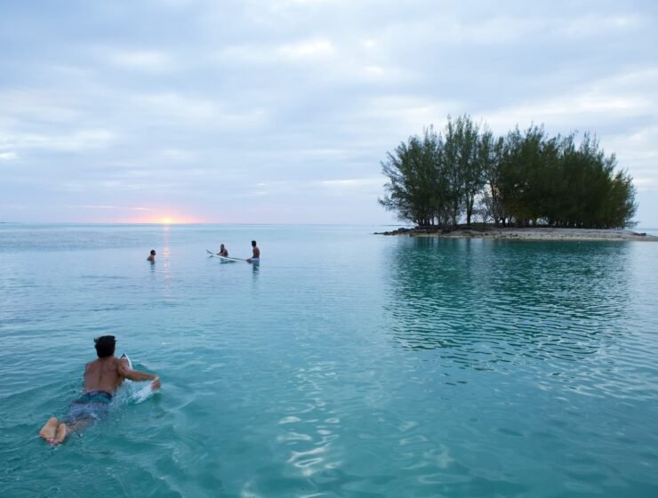 un homme au premier plan dans le lagon de Moorea allongé sur une planche de surf, nageant vers 3 autres personnes en train de discuter, assis sur une planche de surf. Motu et couché de soleil en arrière plan. Le guide ultime des sports nautiques en Polynésie française.