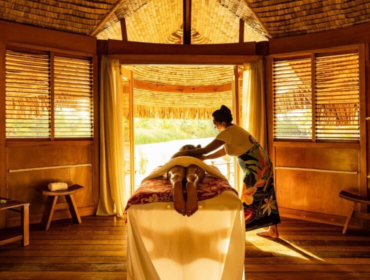 dans un fare traditionnel ouvert sur un lac à Taha'a, une femme se fait masser par une autre femme polynésienne, allongée sur une table. Bien-être à la Polynésienne : Les Secrets de la Santé et de la Relaxation.