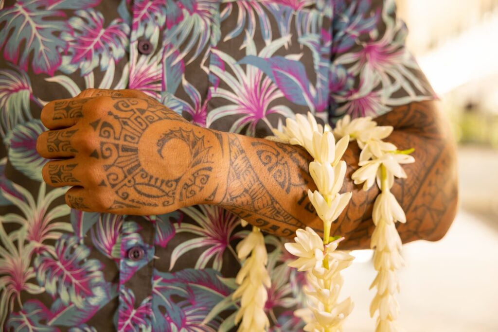 buste avec bras relevé d'un homme polynésien tatoué tenant des collier de fleurs de tiare en guise de bienvenu.