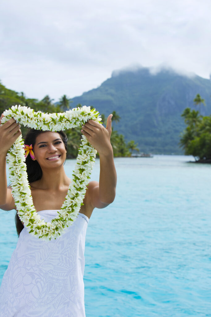 Femme polynésienne tend les bras avec un collier de fleurs de Tiare en signe de bienvenu. Lagon et île de Bora Bora en arrière plan.