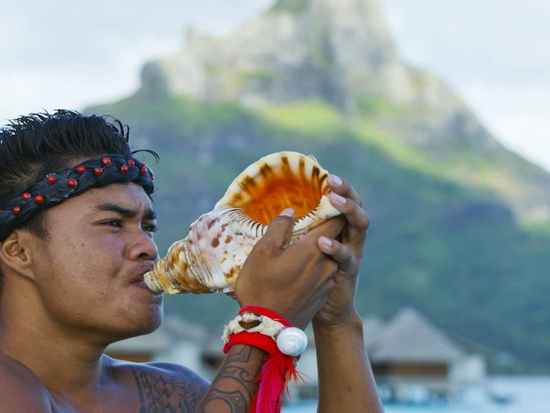 Homme polynésien tatoué soufflant dans un Pu'u en signe de bienvenue. Mont Otemanu flouté en arrière plan.