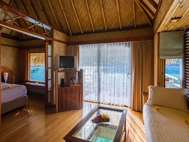 Hôtel Intercontinental Le Moana 4* à Bora Bora - vue de l'intérieur d'un bungalow sur pilotis avec un salon avec table à fond de verre, et une chambre à coucher avec vue sur le lagon.