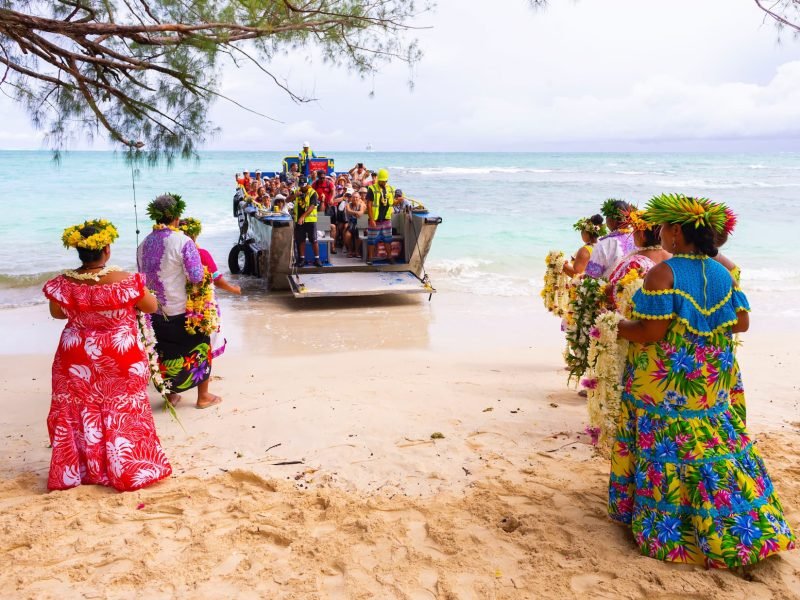 Hommes et femmes polynésiens accueillant sur la plage une barge de l'Aranui avec des voyageurs dedans.