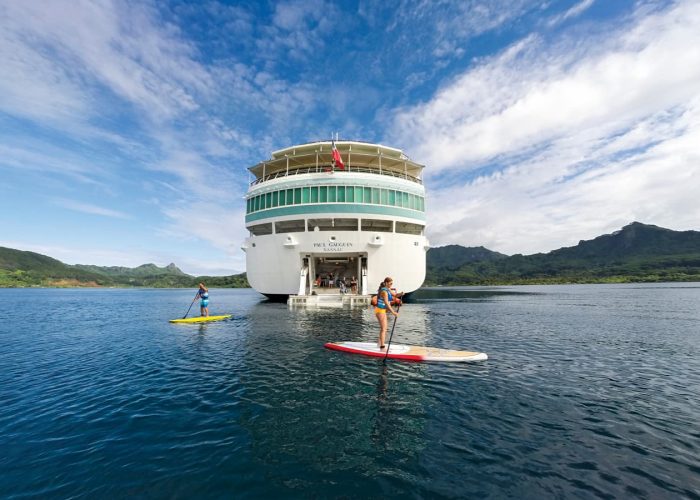 Croisière Paul Gauguin – Ponant. Poupe du paquebot avec 2 touristes faisant du Stand Up Paddle dans le lagon.