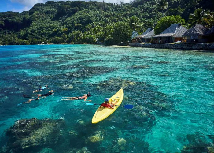 Hôtel Le Mahana 3* à Huahine. Vue depuis le lagon sur l'hôtel et la montagne. 1 personne fait du kayak et 3 autres font du snorkelling au premier plan.