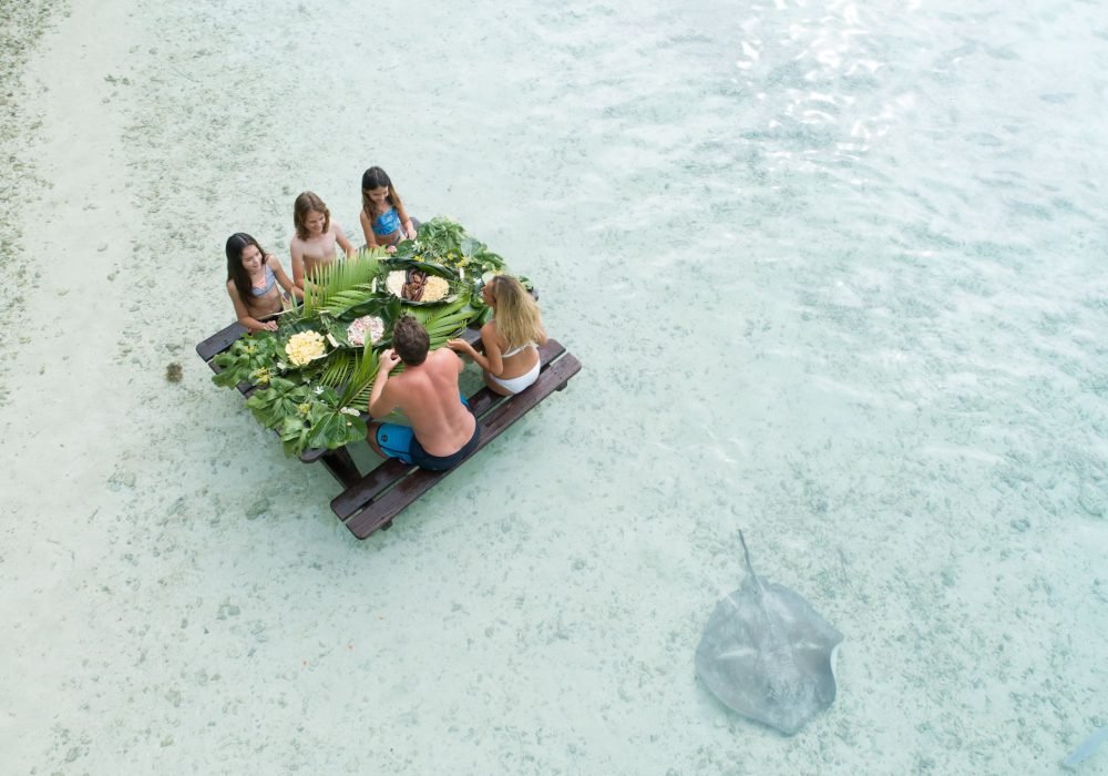 Vue aérienne d'une famille mangeant un repas polynésien les pieds dans l'eau. Une raie pastenague passe à côté d'eux.
