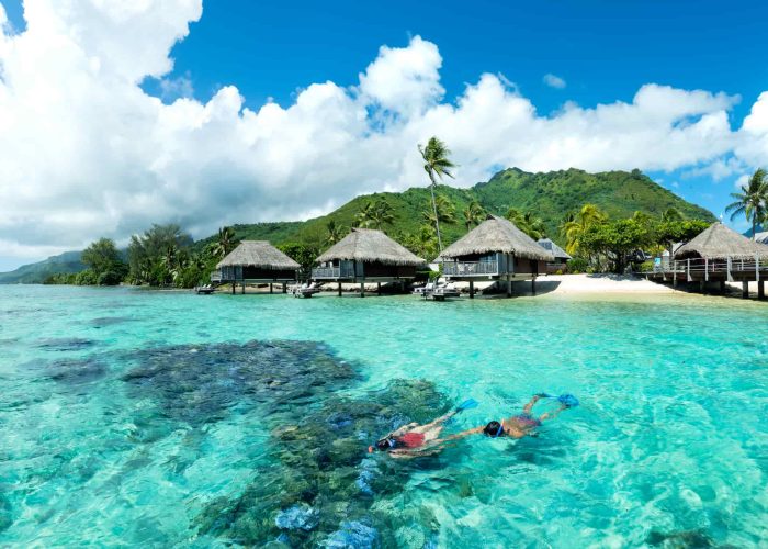 Hôtel Hilton Lagoon Resort & Spa 5* à Moorea. Deux personne faisant sur snorkelling dans le lagon. 3 Bungalows sur pilotis et montagne et arrière plan.