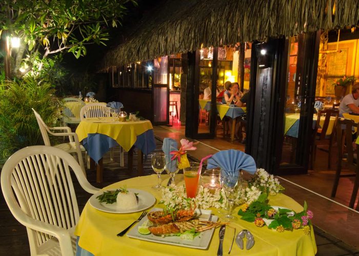 Hôtel Les Tipaniers 2* à Moorea. Restaurant côté Jardin intérieur et extérieur. Table dressée au premier plan avec langoustes et cocktails.