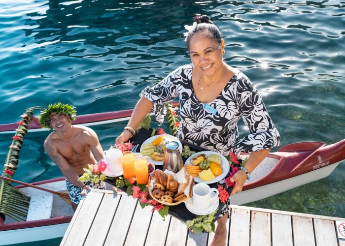 Hôtel Manava Beach Resort & Spa 4* à Moorea. Femme polynésienne apportant un petit déjeuner sur la terrasse d'un bungalow sur pilotis. Homme tahitien et vaa'a en contrebas.