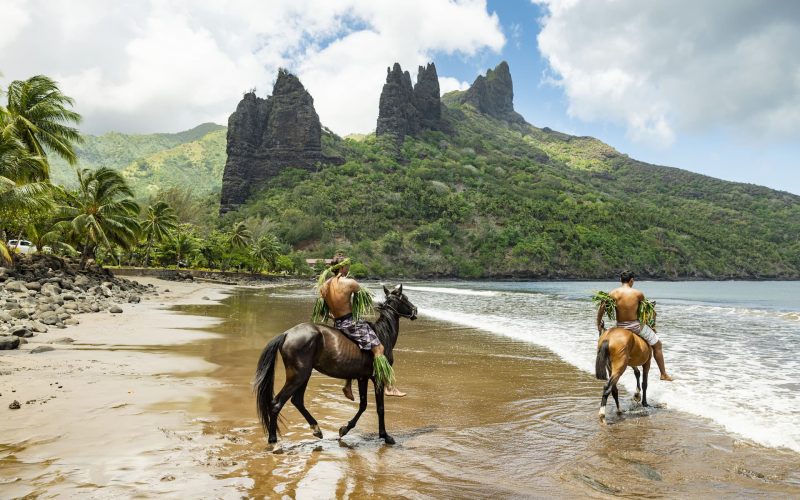 deux marquisiens à cheval sur la plage du village d'Hatiheu à Nuku Hiva. Pics montagneux en arrière plan