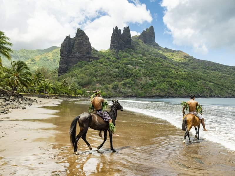 deux marquisiens à cheval sur la plage du village d'Hatiheu à Nuku Hiva. Pics montagneux en arrière plan