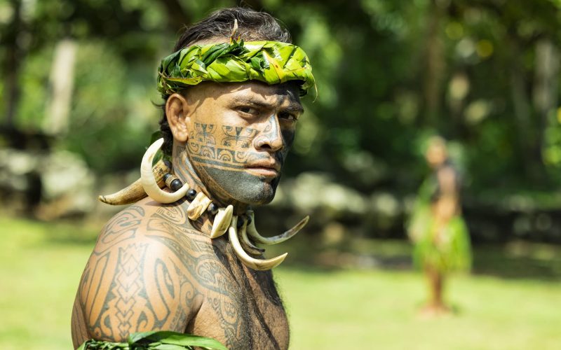 portrait d'un guerrier marquisien, tourné de trois quart. Il est tatoué sur le corps et sur le visage, et il porte un collier fait avec des défenses de cochon