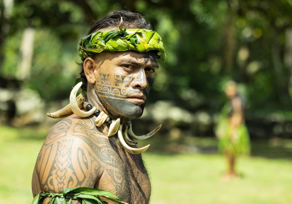 portrait d'un guerrier marquisien, tourné de trois quart. Il est tatoué sur le corps et sur le visage, et il porte un collier fait avec des défenses de cochon