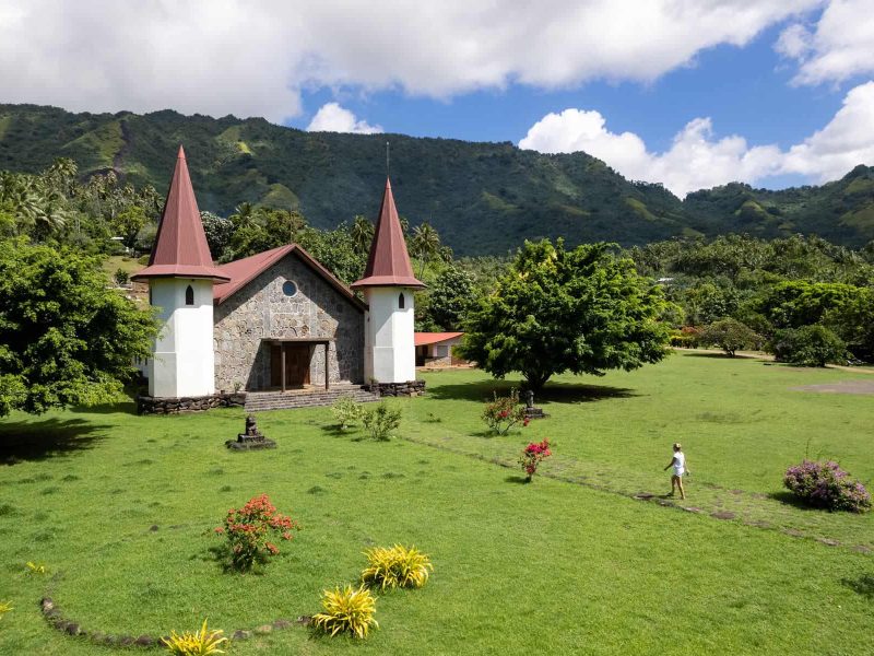 Excursion à Hatiheu organisé par l'hôtel Le Nuku Hiva by Pearl Resorts 3*. Vue du l'église du village d'Hatihey avec une femme marchant en direction de l'entrée.