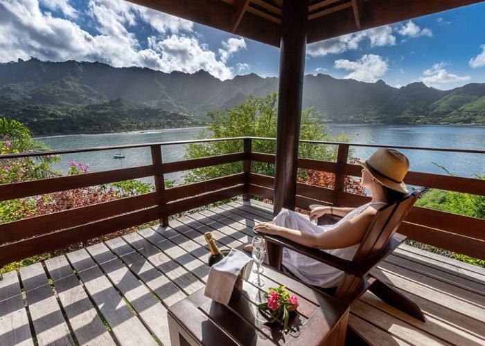 Hôtel Le Nuku Hiva by Pearl Resorts 3* à Nuku Hiva – Femme assise sur une chaise en bois sur la terrasse d'un Bungalow avec vue sur le Baie de Taihoae.