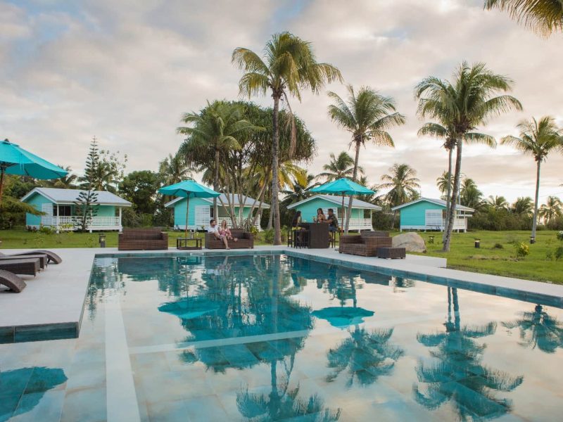 Hôtel Raiatea Lodge 3* à Raiatea - vue sur la piscine au coucher du soleil, avec des personnes sirotant un cocktail. Cocoteraie et bungalows en arrière plan