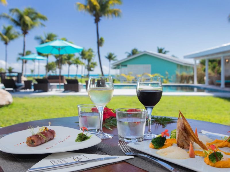 Hôtel Raiatea Lodge 3* à Raiatea - vue sur une table pour deux personnes avec des plats typiques polynésiens et deux verres de vin. Piscine et cocoteraie en arrière plan.