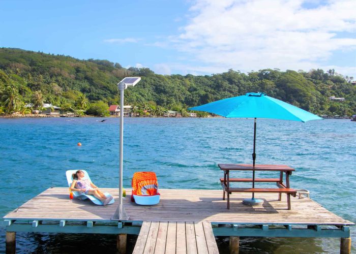 Pension Villa Ixora à Raiatea. Ponton de la pension avec une femme prenant urbain de soleil sur un transat. Table de pique nique et parasol.