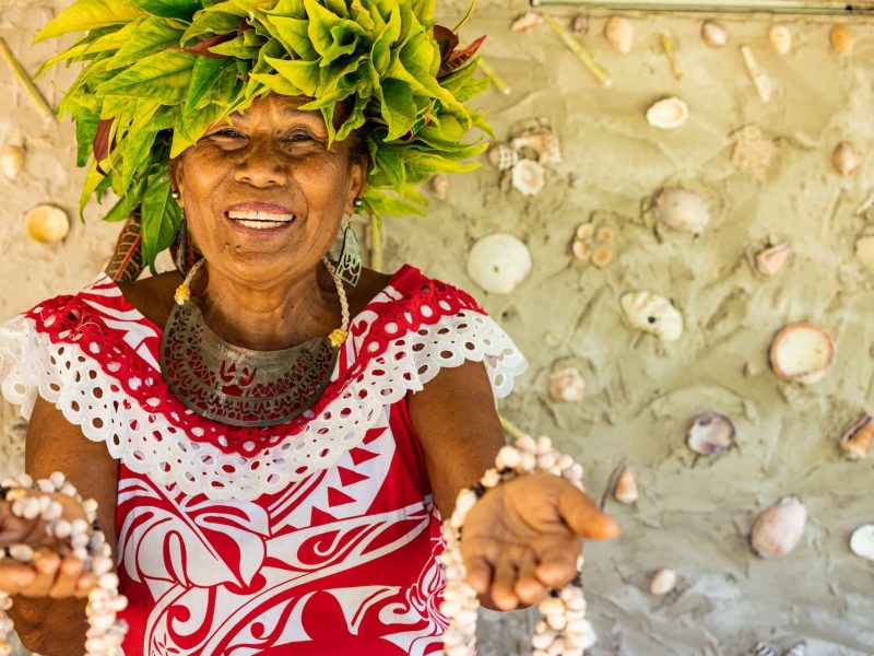 Femme polynésienne avec une couronne végétale sur la tête, sourit et tends les mains en avant avec des colliers en coquillages en signe de bienvenue