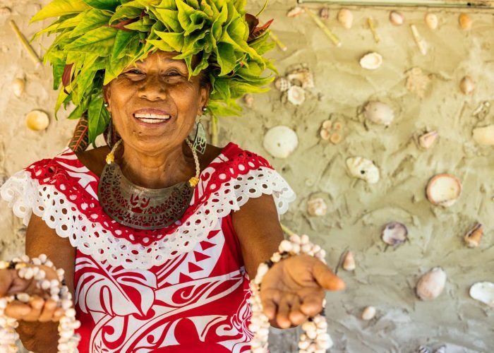 Femme polynésienne avec une couronne végétale sur la tête, sourit et tends les mains en avant avec des colliers en coquillages en signe de bienvenue