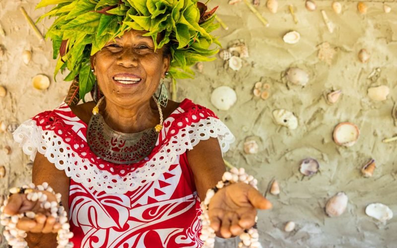 Femme polynésienne avec une couronne végétale sur la tête, sourit et tends les mains en avant avec des colliers en coquillages en signe de bienvenue