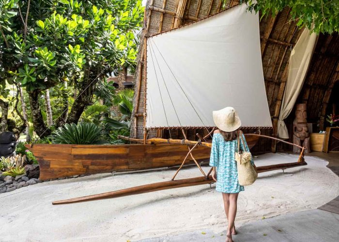 Hôtel Le Taha’a by Pearl Resorts 5*. Femme portant un chapeau marchant vers l'entrée de la réception et sa grande pirogue à balancier.