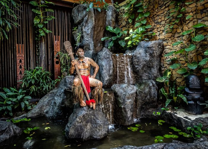 Hôtel Le Taha’a by Pearl Resorts 5*. Homme Polynésien tatoué de la tête au pieds assis dans la cascade de la réception de l'hôtel.