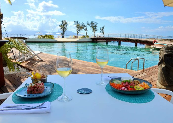 Hôtel Te Moana 3* à Tahiti - vue d'une table avec deux entrées et verres de vin. Piscine et lagon en deuxième plan