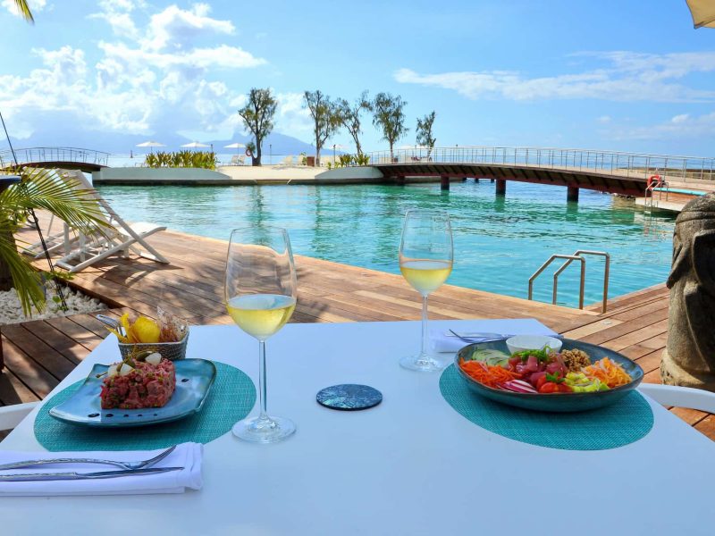 Hôtel Te Moana 3* à Tahiti - vue d'une table avec deux entrées et verres de vin. Piscine et lagon en deuxième plan