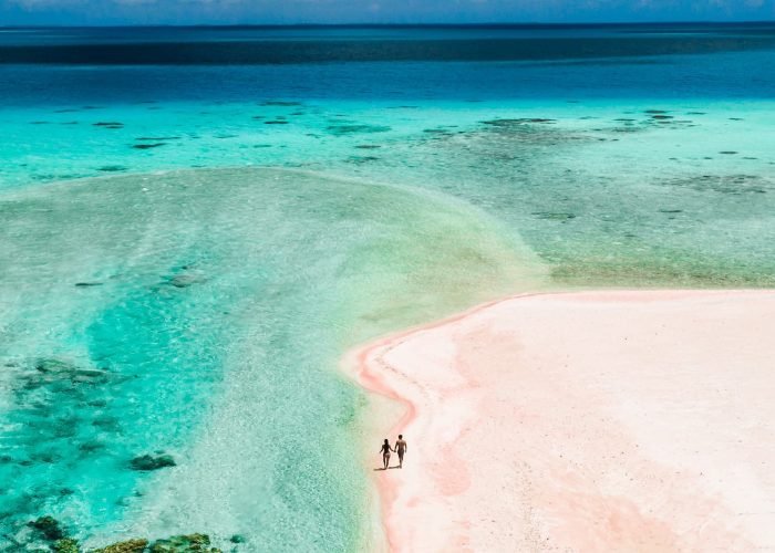 subtiles-tahiti-tikehau-hôtel-le-tikehau-by-pearl-resorts-2©le-tikehau-by-pearl-resorts Hôtel Le Tikehau by Pearl Resorts 4*- vue aérienne d'un couple marchant sur une plage de sable rose, au bord d'un lagon turquoise.
