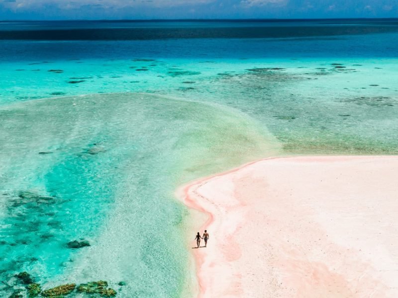 Hôtel Le Tikehau by Pearl Resorts 4*- vue aérienne d'un couple marchant sur une plage de sable rose, au bord d'un lagon turquoise.