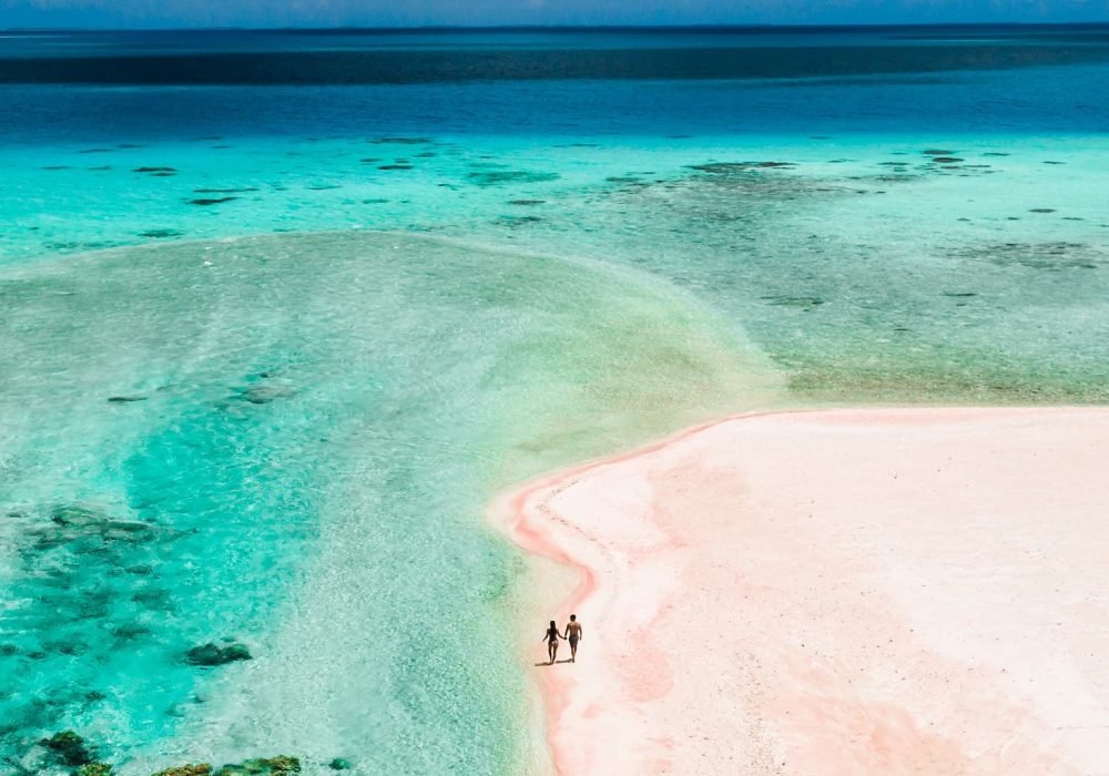 Hôtel Le Tikehau by Pearl Resorts 4*- vue aérienne d'un couple marchant sur une plage de sable rose, au bord d'un lagon turquoise.